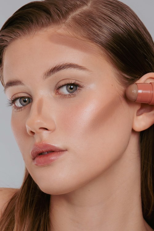 A woman applies makeup or skincare to her face, looking at the camera with a neutral expression.