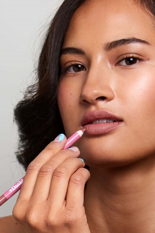 Een vrouw brengt lippenstift aan met haar vinger, ziet er rustig uit met natuurlijke make-up en donker haar. De achtergrond is eenvoudig.
