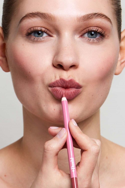 A woman with blue eyes and fair skin is applying pink lipstick, holding the lipstick close to her lips.