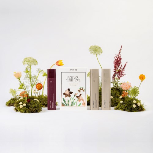 Fyra flaskor av parfym eller lotion omges av blommor och mossa, vilket skapar en frisk och naturlig presentation.