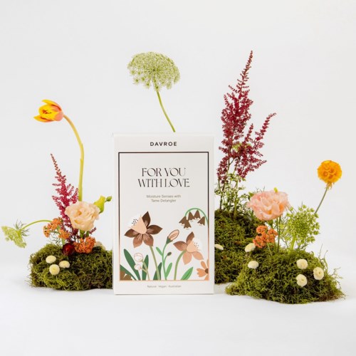 En blomsteruppsättning med färgglada blommor och grön mossa, centrerad runt ett kort som säger "FOR YOU WITH LOVE."
