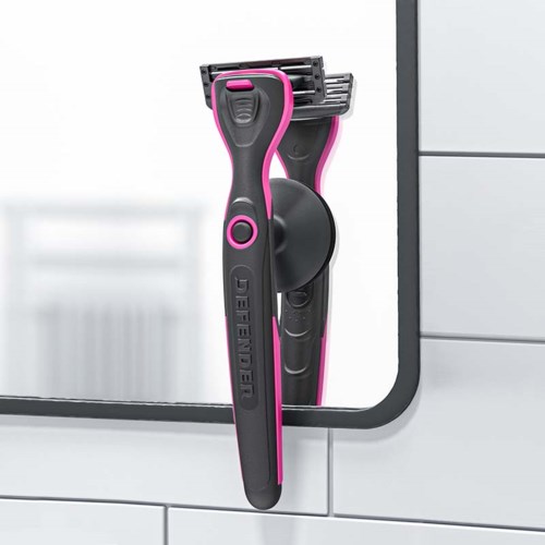 A pink and black razor is attached to a mirror in a bathroom. The mirror is part of a white tiled wall.