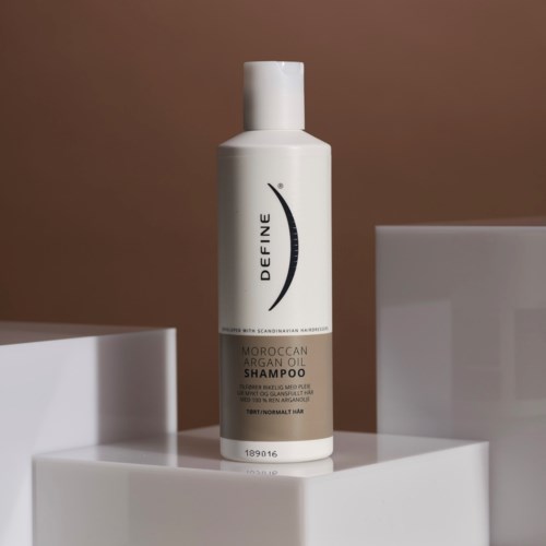 A white bottle of Moroccan argan oil shampoo on a white platform, with a brown background.