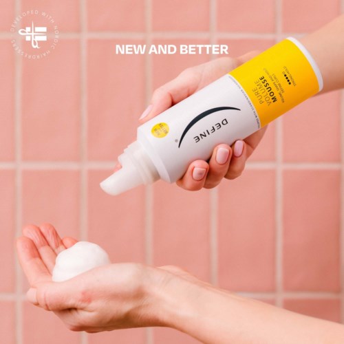 A person dispenses foam from a yellow and white skincare bottle onto their hand in front of pink tiles.