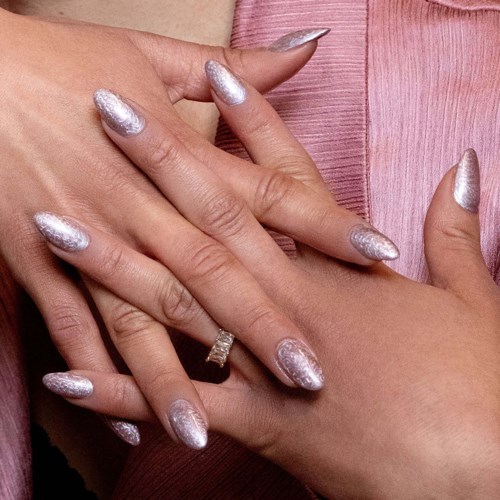 Two hands with silver glittery nails, one hand showing a ring, against pink fabric.