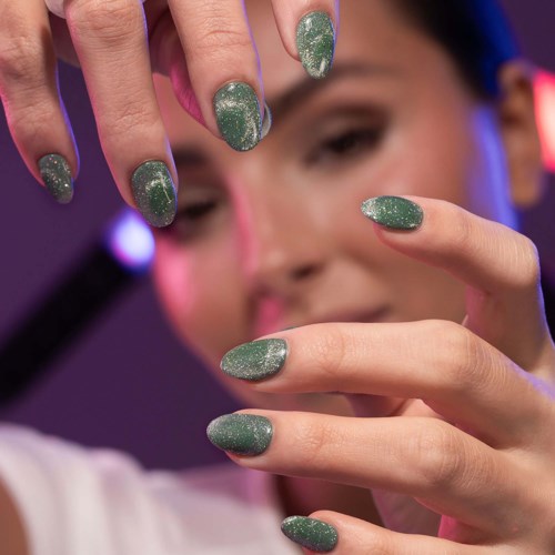 A person shows four fingers with glittery green nail polish. The background has vibrant pink and purple lights.