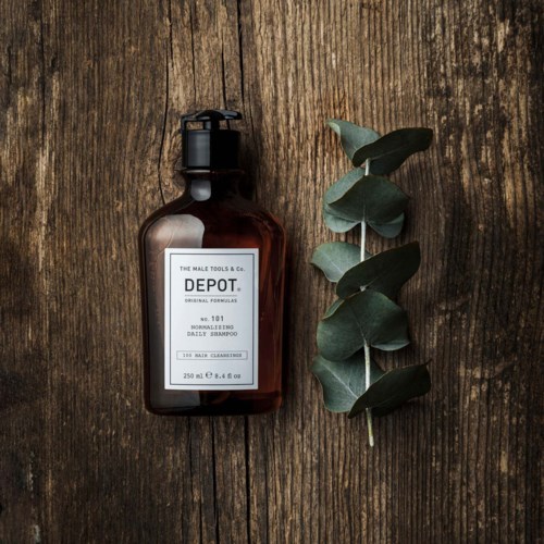 A brown shampoo bottle with a black pump lies on wooden surface next to a eucalyptus branch.