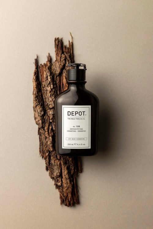 Een zwarte fles met label 'DEPOT' bevestigd aan een stuk ruwe schors, met een eenvoudige achtergrond.