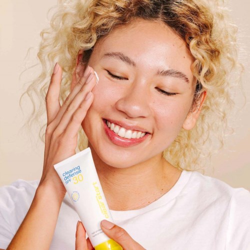 Een lachende vrouw met krullend blond haar smeert zonnebrandcrème op haar gezicht.