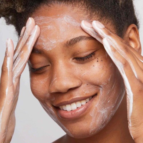 A woman is washing her face and smiling, with soap or cleanser on her face and hands.