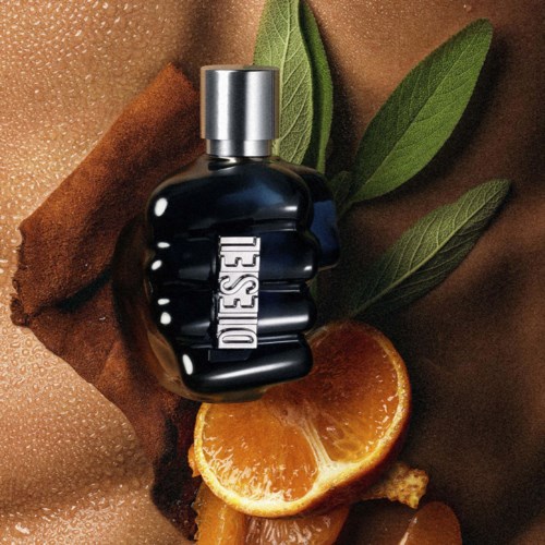 A Diesel perfume bottle is surrounded by cinnamon, orange slices, leaves, and a textured background.