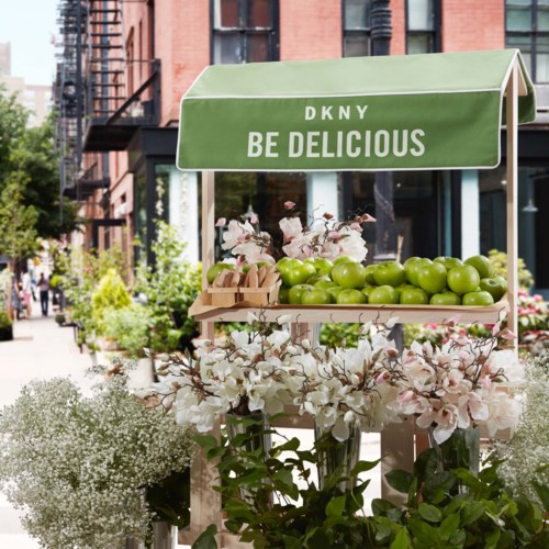 Een fruitkraam met groene appels en roze bloemen, onder een bord met "Be Delicious," in een stedelijke straat.