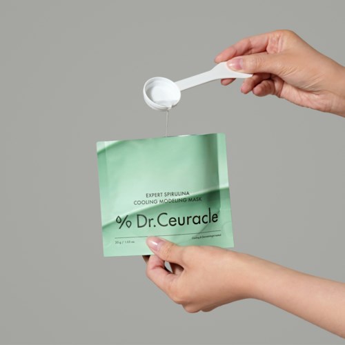 Iemand houdt een groene gezichtsmasker vast en een lepel schept witte crème of gel uit een klein container.