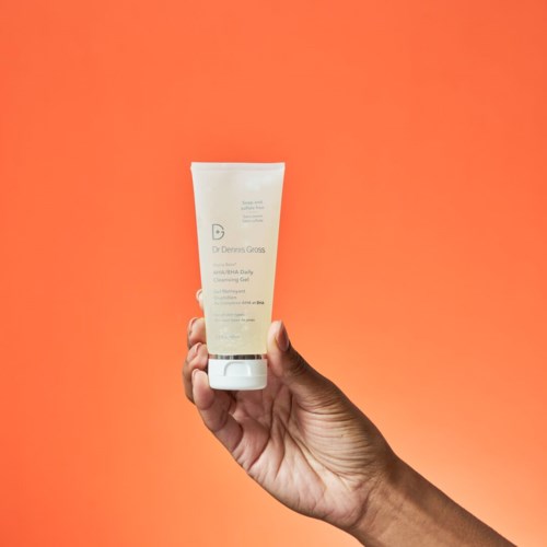 Eine Hand hält eine weiße Tube mit Reinigungs Gel vor orangefarbenem Hintergrund.