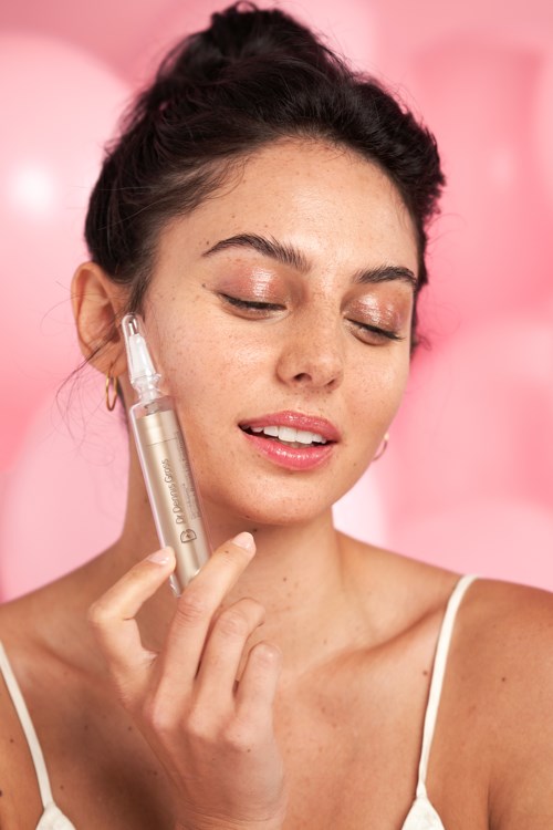 Eine Frau mit geschlossenen Augen trägt Serum auf ihr Gesicht auf vor rosa Hintergrund.