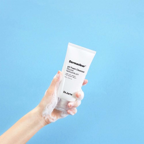 En hand håller ett vitt rör märkt "Dermaclear pH Foam Cleanser" mot en ljusblå bakgrund.