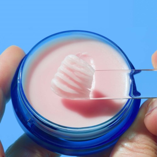 A hand holding a small jar of pink cream with a spatula inside, set against a blue background.