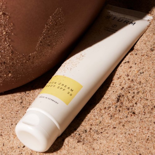 Eine weiße Tube mit Sonnencreme SPF 30 liegt im Sand neben einem Arm.