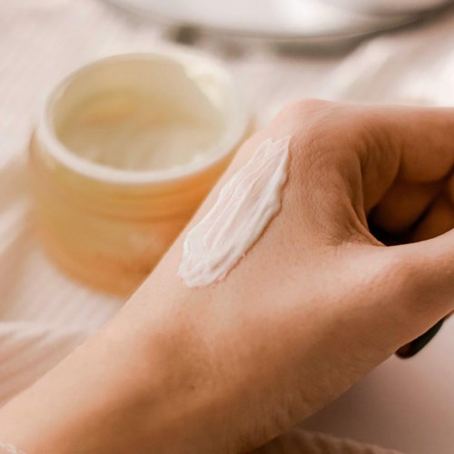 Een hand met crème erop, naast een open pot crème op een witte ondergrond. Een lepel is zichtbaar op de achtergrond.
