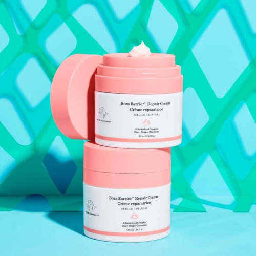 To rosa beholdere med Bora Barrier Repair Cream stablet mot en lys blå bakgrunn med et grønt mønster.
