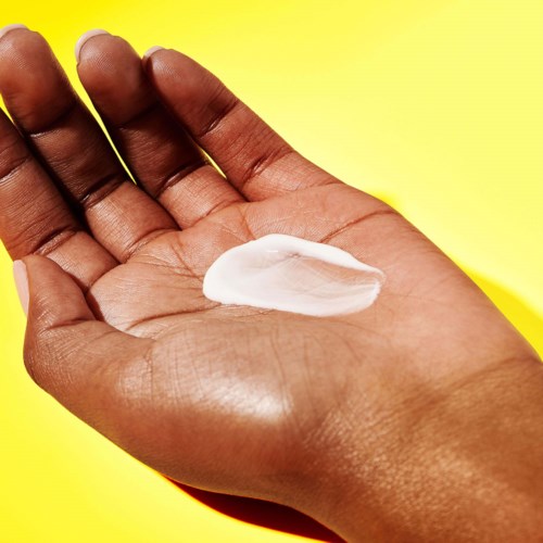 En hånd holder en hvit lotion eller krem på en lys gul bakgrunn.