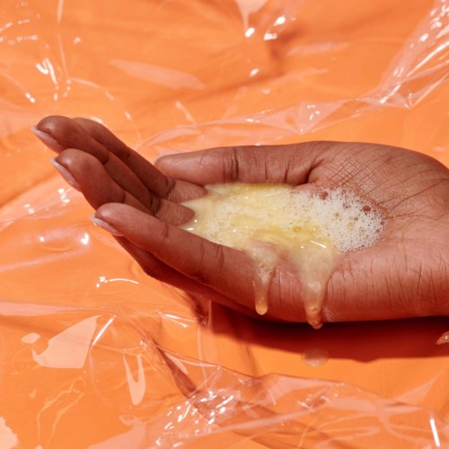En hånd dytter ut en gul væske i en gjennomsiktig plasticpose på en oransje overflate.
