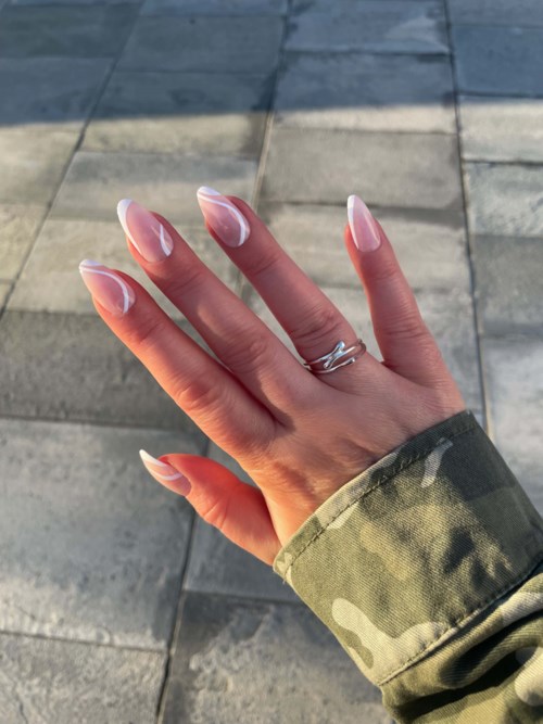 A hand with long, neatly shaped nails is shown, wearing a camouflage jacket against a paved ground.