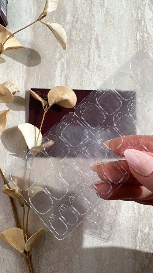 A hand peeling off clear sticker sheets with nail designs, next to a potted plant and a closed box.
