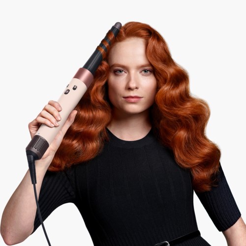 A woman with long, curly red hair uses a curling iron on her hair against a white background.