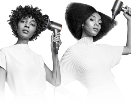 Two women with big curly hair styling their hair with hairdryers, in a black and white photo.