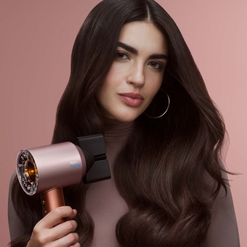 A woman using a hair dryer, with long, dark, shiny hair and a neutral pink background.