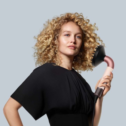 A woman with curly blonde hair holding a hairbrush hairdryer. She wears a black outfit and looks confidently.