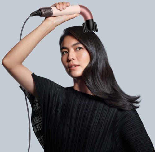 A woman with long black hair using a pink hair straightener on her head.