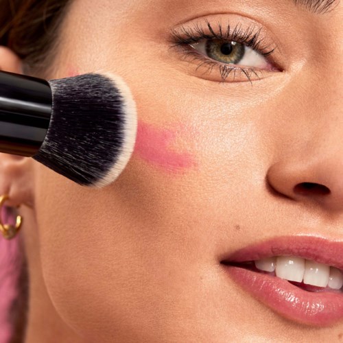 A woman is blush on her cheek with a makeup brush. She is smiling and has earrings and makeup.