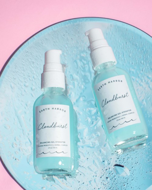 Two spray bottles labeled 'Cloudburst' placed on a blue dish, with water droplets and pink background.