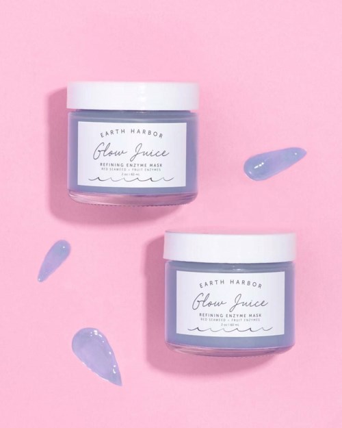 Zwei Gläser Earth Harbor Glow Juice Enzymmaske auf rosa Hintergrund, mit lila Cremestücken drumherum.