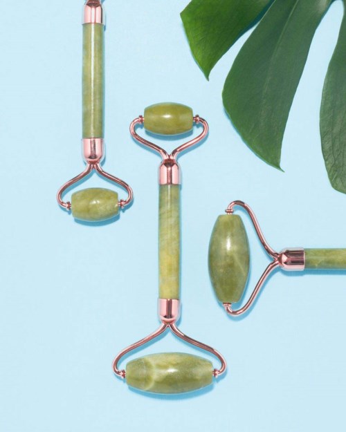 Gröna jade ansiktsrollers med roségolddetaljer, placerade på en ljusblå yta nära gröna blad.