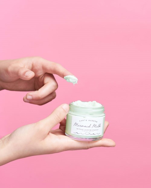 Zwei Hände halten ein Glas mit grüner Creme vor rosa Hintergrund. Eine Hand schöpft Creme mit einem Finger.