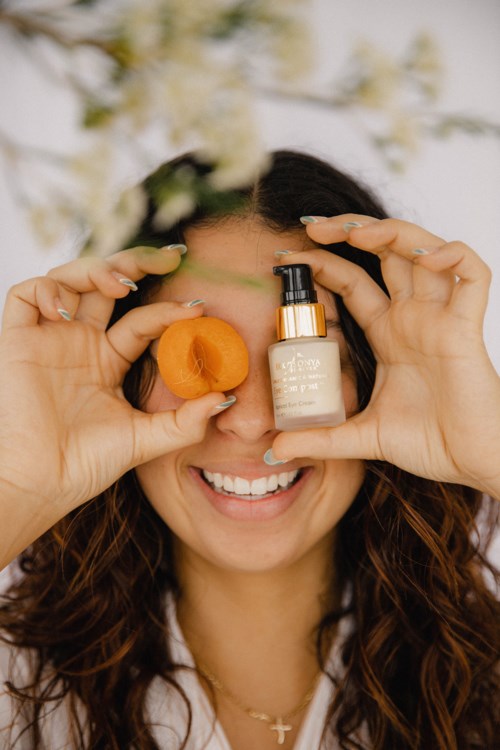 A woman smiles while holding a small orange fruit and a bottle of skincare product in front of her face.