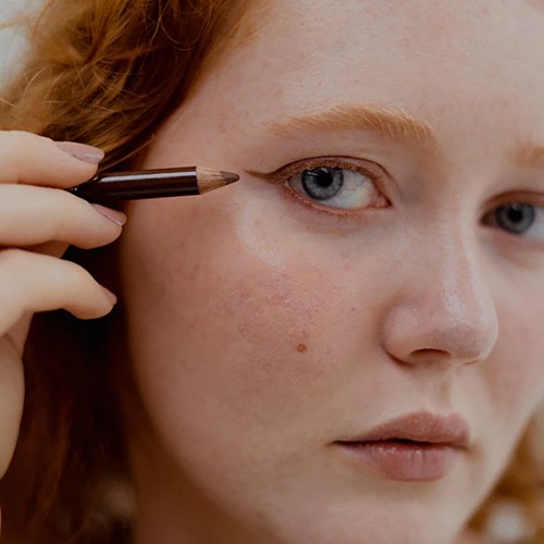 Eine junge Frau trägt Make-up in der Nähe ihres Auges mit einem braunen Stift oder Pinsel auf.