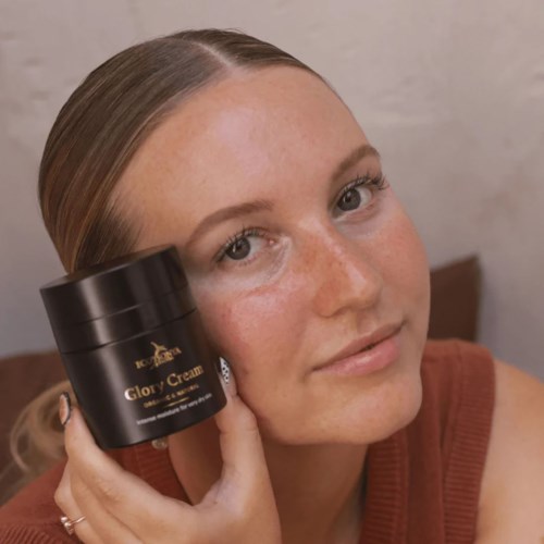 Een vrouw met een heldere huid houdt een zwarte pot met Glory Cream dicht bij haar gezicht.