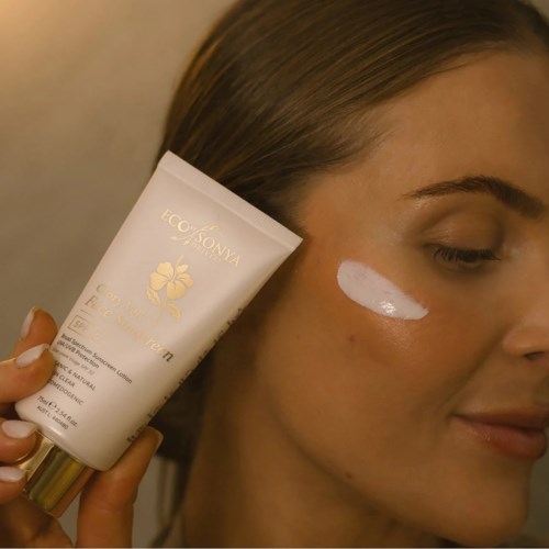 Een vrouw met een glimlach brengt witte crème aan onder haar oog. Ze houdt een tube vast met het label Ecoc Sonya. De achtergrond is eenvoudig.