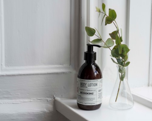Eine braune Flasche mit 'Body Lotion' Beschriftung neben einer kleinen Glasvase mit grünen Blättern auf einem Fensterbrett.