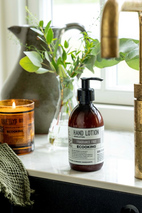 A brown bottle of hand lotion with a pump sits on a windowsill, next to a candle and green plants.
