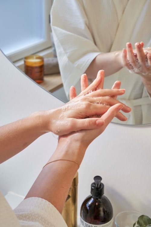 Person washing hands in front of a mirror, soap and bathroom items visible.
