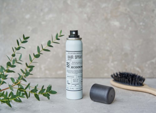 A white bottle of hair spray, a black cap, a wooden brush, and green plant leaves on a light surface.