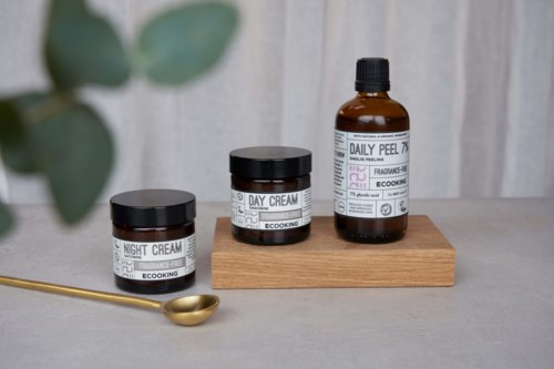 Three skincare jars and a bottle on a wooden stand, with a gold spoon nearby, set on a gray surface.