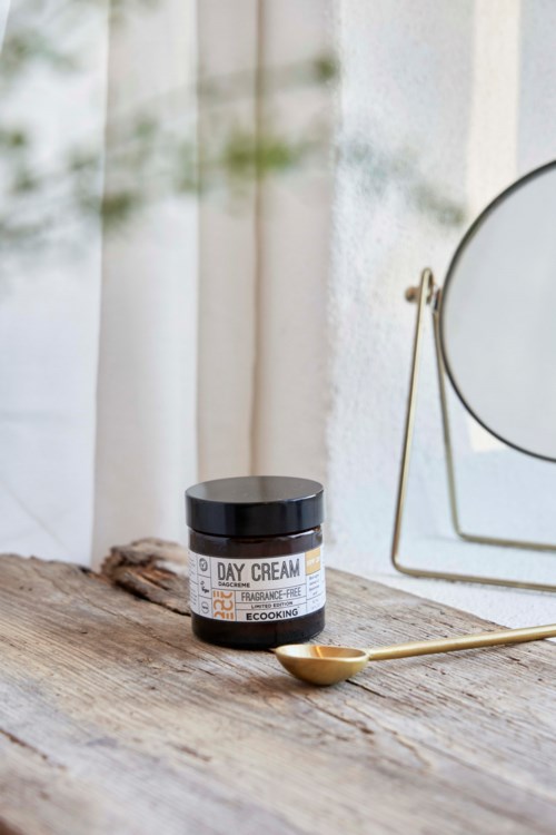 A jar of day cream on a wooden surface with a spoon and a mirror nearby.