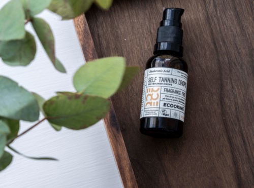 A small dark bottle labeled as self-tanning drops sits on a wooden surface next to green leaves.
