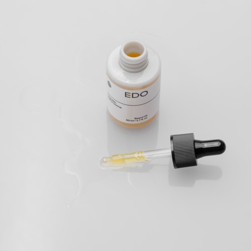 Kleine Flasche mit der Aufschrift 'EDO' und Pipette mit orangefarbener Flüssigkeit. Flasche liegt offen auf weißer Oberfläche.